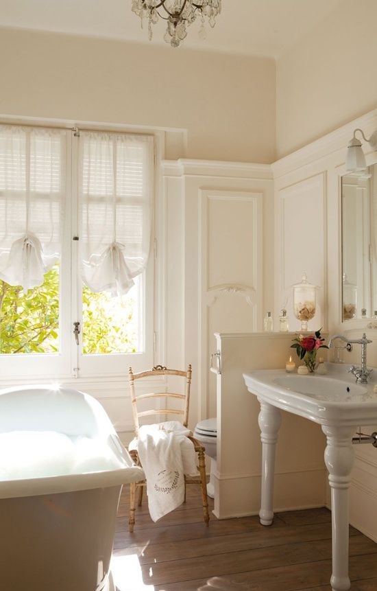 a light-filled farmhouse bathroom done in creamy shades, a clawfoot bathtub, elegant curtains-shades, a vintage vanity