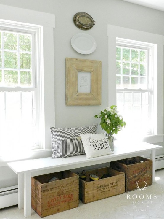 a neutral farmhouse entryway with an artwork, decorative plates, a bench, wooden boxes for storage and a striped pillow