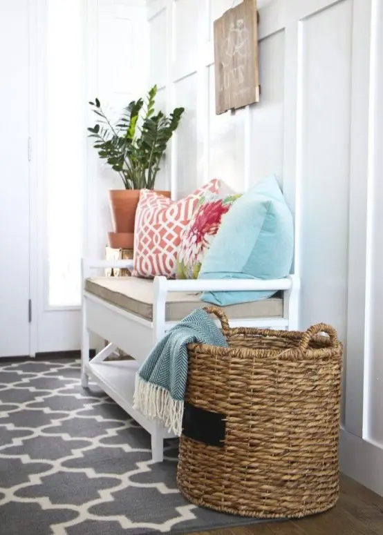 a cozy farmhouse entryway with a vintage white bench with colorful pillows, a wooden sign and a basket with blankets