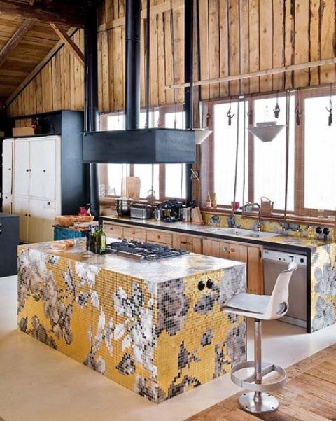 A unique chalet kitchen clad with light colored wood, dark metal appliances and colorful yellow and grey tiles forming mosaics on all the countertops