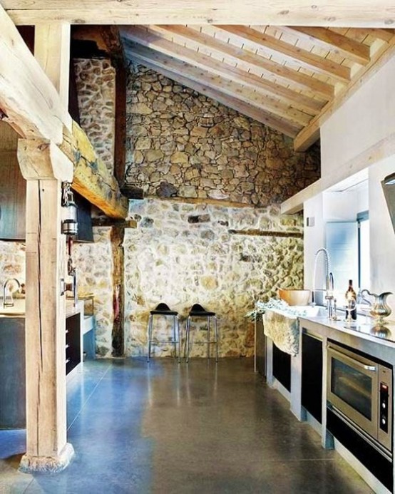 a chalet kitchen done with stone walls, with a wooden ceiling with beams and wooden beams right in the space is a cool idea for a chalet feel