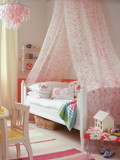 a lovely girl's room with neutral walls and a floor, a white bed with floral bedding, a pink floral canopy and a wall-mounted shelf, lovely toys and chairs