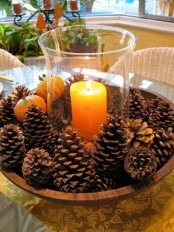 a simple and eays to make fall centerpiece of a wooden bowl with pinecones, faux pumpkins and a candle in a clear candle holder