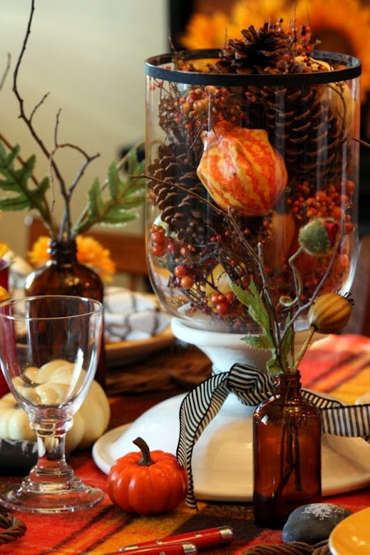 a simple fall centerpiece of a tall clear vase filled with fake berries, pinecones and gourds