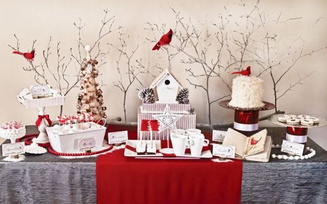 A catchy Christmas sweets table with a grey tablecloth, red runners and beads, white sweets and mugs and some red birds sitting on the branches over the table