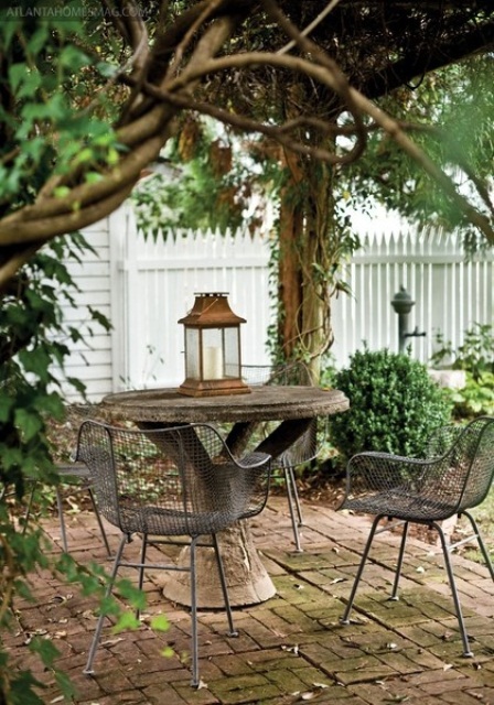 a mini rustic pergola all covered with plants, with a stone table and metal chairs