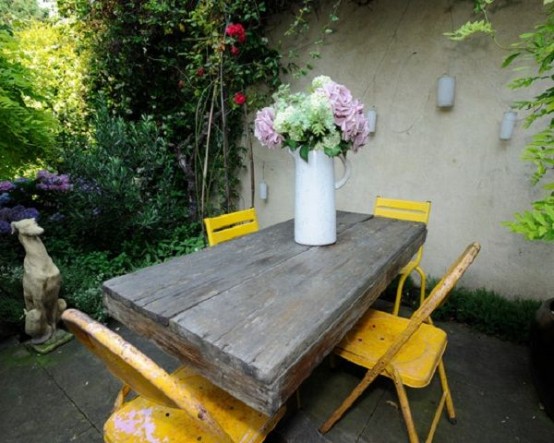 a cozy rustic patio with a wooden table, bold yellow chairs and potted blooms