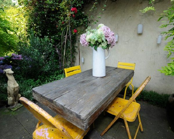 a cozy rustic patio with a wooden table, bold yellow chairs and potted blooms
