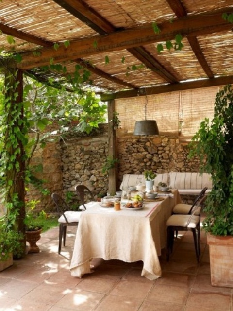 a welcoming patio with stone walls, wooden furniture, potted greenery and wicker lampshades