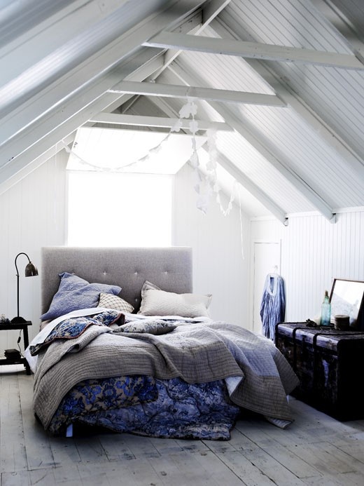 A light filled Nordic bedroom with white planks and beams on the ceiling, a grey uphosltred bed and bright bedding