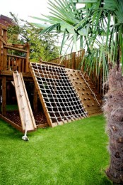 a small outdoor playground with green lawn, ladders, nets, a small tree house and a sand box under it