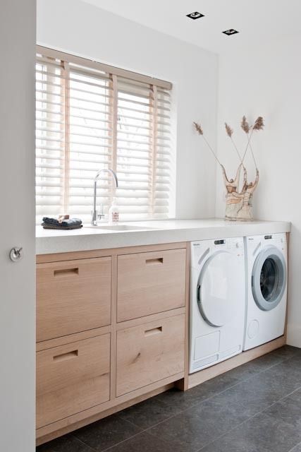 A neutral laundry with sleek light stained drawers, a concrete countertop, a washing machine and dryer and some grasses
