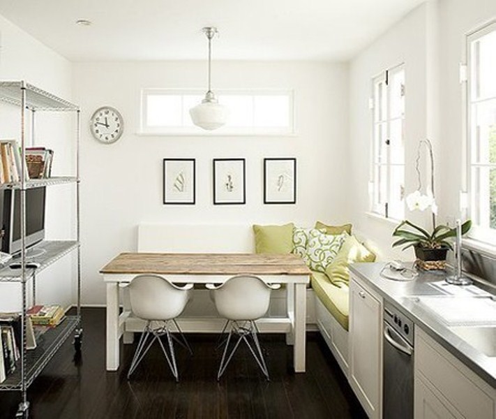 A small contemporary kitchen in white, with an L shaped bench, a wooden top table, a large open storage unit