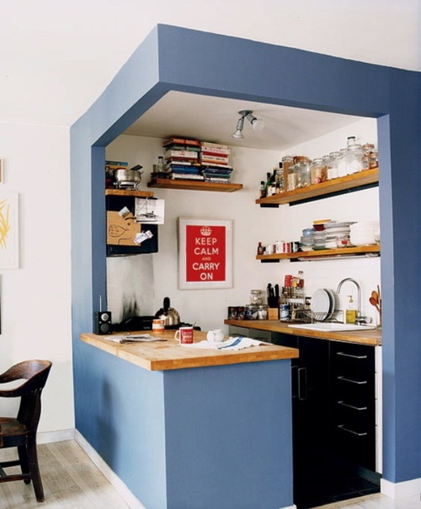 A small kitchen placed in a blue cube, with built in black cabinets, light colored woodne countertops and open shelving