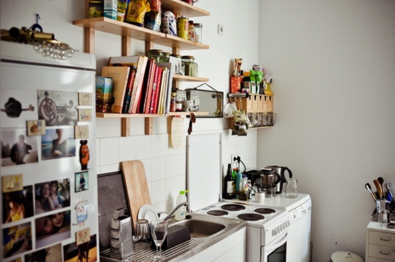 A small contemporary kitchen in white, with light stained wooden shelves and lots of personal photos