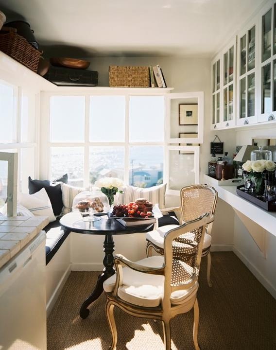 A small farmhouse kitchen with a view, an L shaped bench, a round table, vintage chairs, glass cabinets and shelves