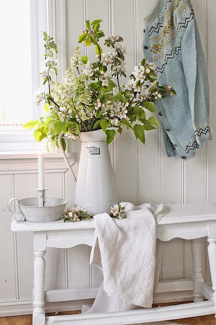 a white shabby chic entryway with a carved bench, a jug with green branches and some blankets