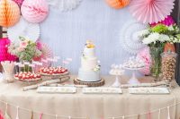 a colorful pink and fuchsia dessert table with paper pompoms and fans, colorful floral arrangements and a bright tassel garland