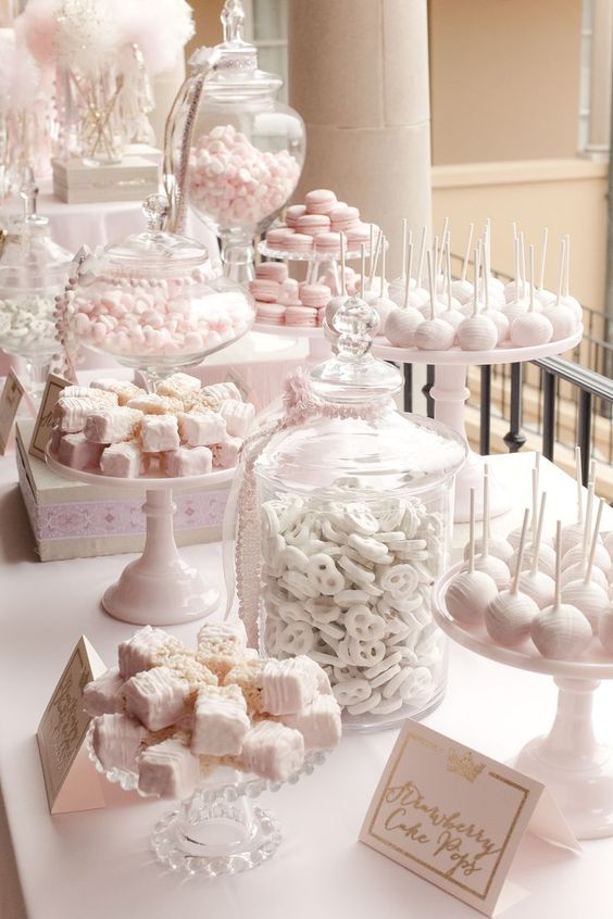 a light pink and white dessert table styled with lots of stands, jars and some signs to mark the sweets