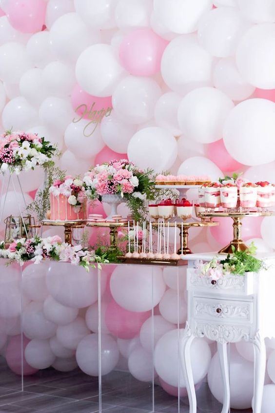 a girlish dessert table with a pink and white balloon wall, bright pink and white floral arrangements, pink desserts and a drip cake