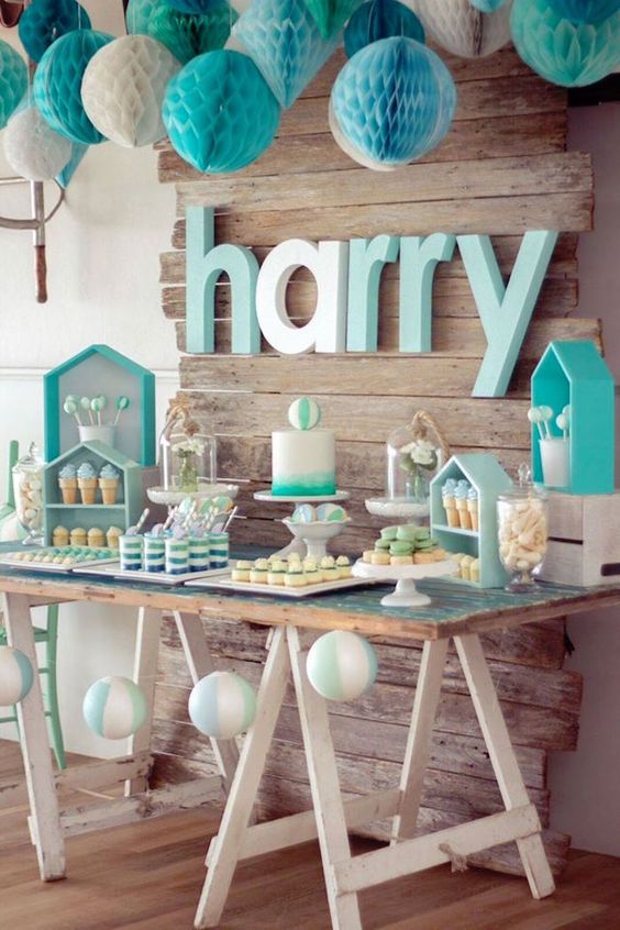 A turquoise and white baby shower dessert table with paper pompoms, house shaped shelves and a trestle table