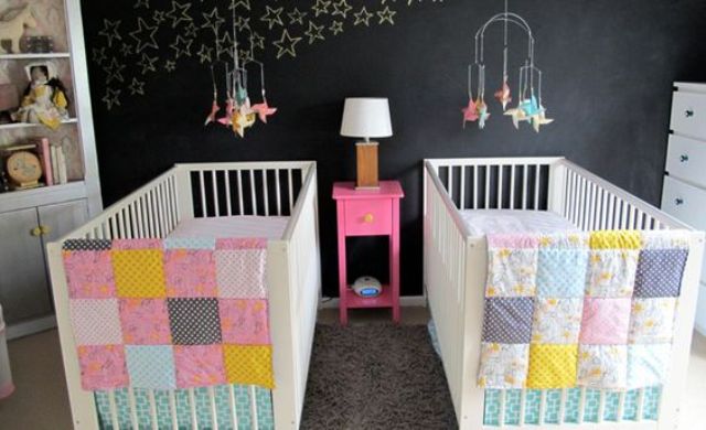 a stylish shared nursery with a chalkboard wall, stars chalked on it, white furniture and colorful patchwork bedding plus colorful mobiles