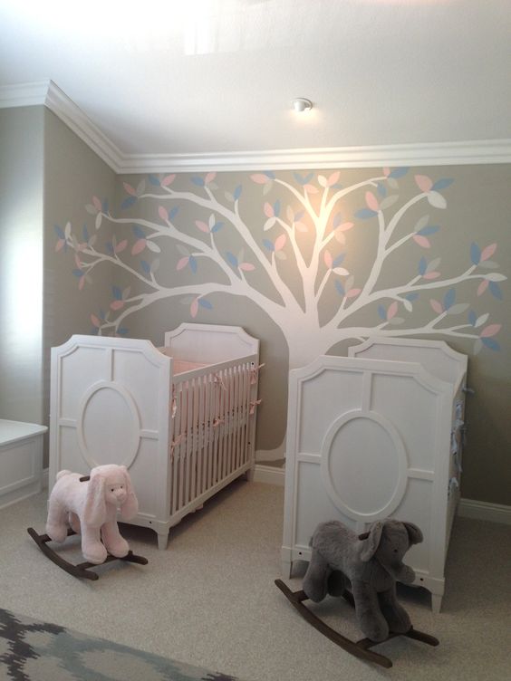 a neutral shared nursery with white furniture, a tree painted on the wall and some toys