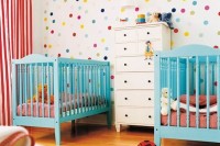 a colorful shared nursery with a bright polka dot wall, turquoise cribes, pink and white bedding and lots of toys