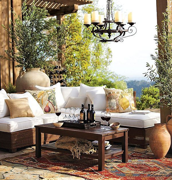 A Mediterranean otudoor space with a white corner sofa, a dark stained coffee table, a vintage candle chandelier, greenery and vases around plus printed pillows