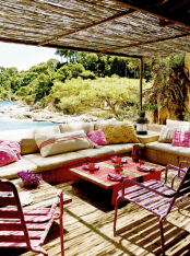 a bright Mediterranean outdoor space with a built-in upholstered bench, low tables, pink pillows, pink metal chairs and a beautiful view