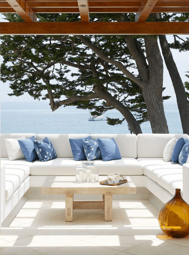 A lovely Mediterranean outdoor space with a built in sofa with white upholstery, blue pillows, a low table and an amber bottle plus a gorgeous sea view