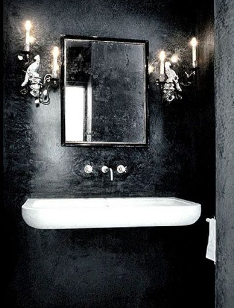 A beautiful Gothic bathroom with textural black walls, a floating white sink, candlelabra styled lamps and a mirror in a modern black frame