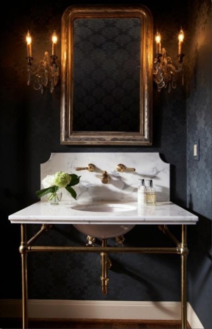 A vintage Gothic bathroom with black printed wallpaper, a free standing sink of white marble, candelabra style lamps and a mirror in a gilded frame