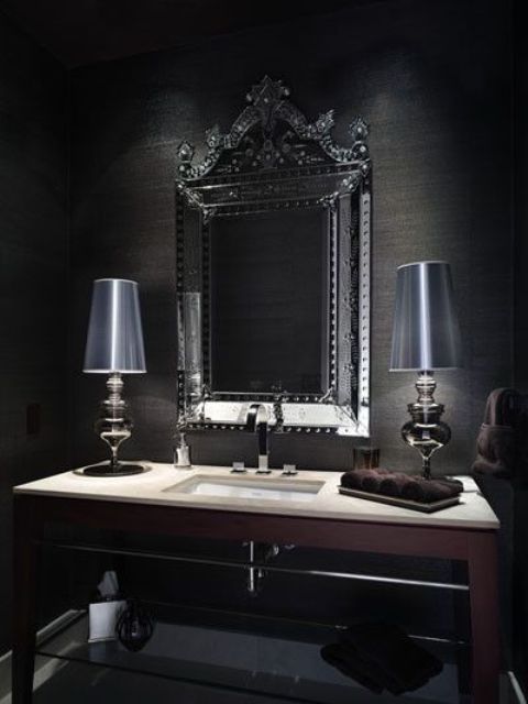 A Gothic bathroom with black walls, a mirror with an ornated frame, chic table lamps and a built in sink