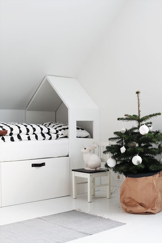 A white Scandi kid's room with a white house shaped bed with drawers, a Christmas tree in a basket and a small nightstand plus printed bedding