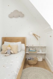 a delicate and subtle Scandinavian kid’s room with a light-stained bed and neutral bedding, an open shelf nightstand, a cloud decoration and a white sconce and pastel bedding