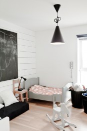 a welcoming Scandinavian kid’s room with a white shiplap wall, a grey bed with pastel bedding, black baskets for storage, a chalkboard and a low sofa with pillows