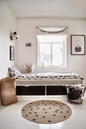 a black and white Scandinavian kid’s room with a white storage bed with black and white bedding, a jute rug and a metal basket for storage, black and white artwork and much natural light