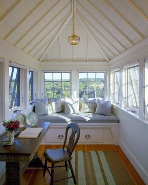 A small attic sunroom with a built in daybed with storage, lots of pillows, a desk and a vintage chair plus a striped rug