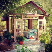 a shabby chic garden shed turned into an outdoor bedroom with a bed with bright bedding, blooms, a rug and lots of greenery around