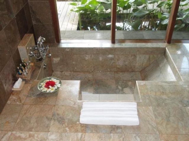 A natural looking bathroom with neutral stone tiles and a sunken bathtub with steps and a glazed wall with a view