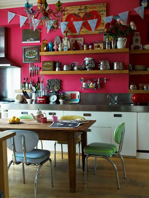 an eclectic kitchen with hot pink walls, stainless steel appliances and a backsplash, a wooden table and pastel chairs
