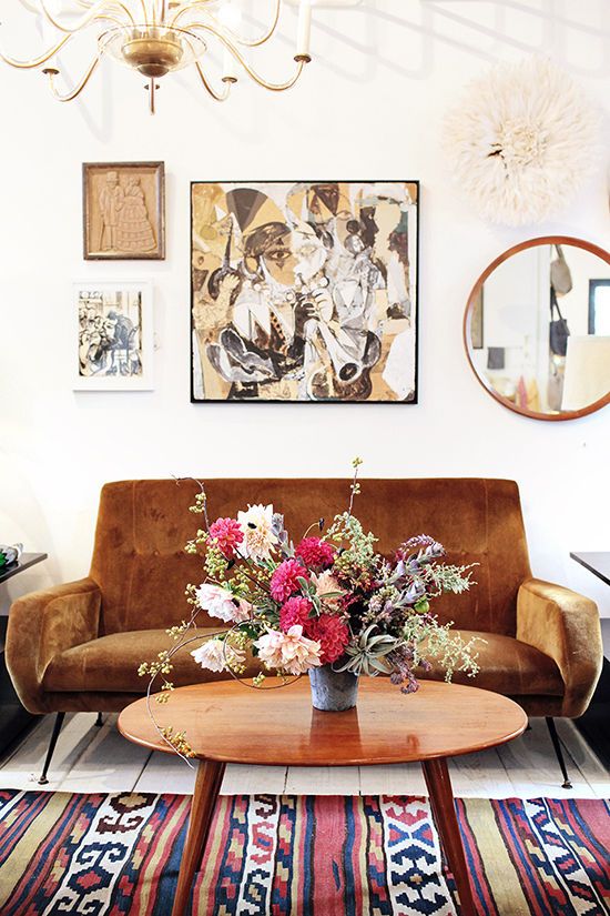 A boho mid century modern space with a mustard mid century modern loveseat, a bright printed rug, a stained table and a gallery wall with a round mirror plus a chic chandelier