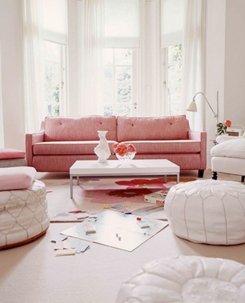 A neutral living room with a bay window, a pink mid century modern sofa, white Moroccan poufs and a chair plus a coffee table