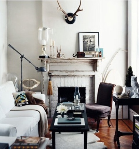 A small eclectic living room with a non workign fireplace, vintage furniture, antlers, artworks and lamps and candles