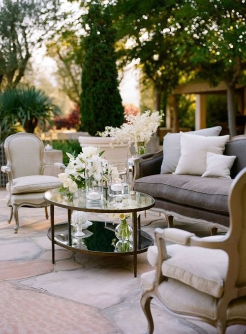 a refined neutral terrace with vintage furniture with neutral upholstery, a tiered glass coffee table and white blooms