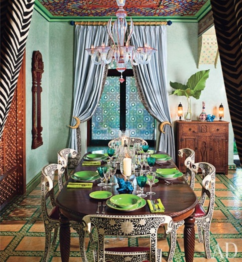 A bright Moroccan inspired dining room with a bright checked floor, a bold ceiling, a heavy carved table and painted chairs plus bold printed textiles