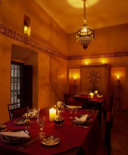 a beautiful Moroccan dining room with tables covered in red, with a refined Moroccan lamp hanging over the space, a pretty patterned door and lovely lamps and candles