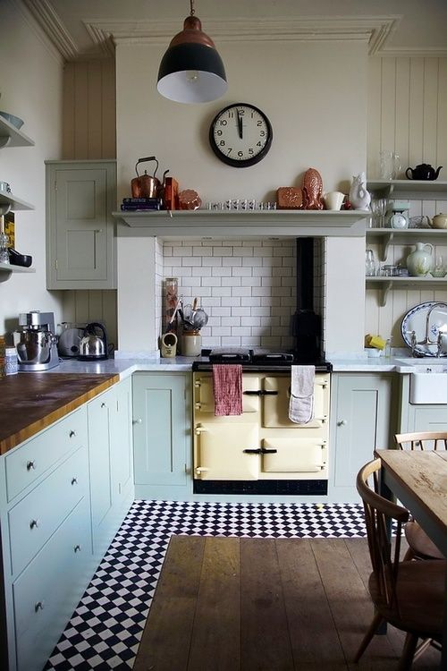 a vintage kitchen with blue cabinets, butcherblock and stone countertops, a vintage cooker, a clock and a pendant lamp plus a black and white checked floor