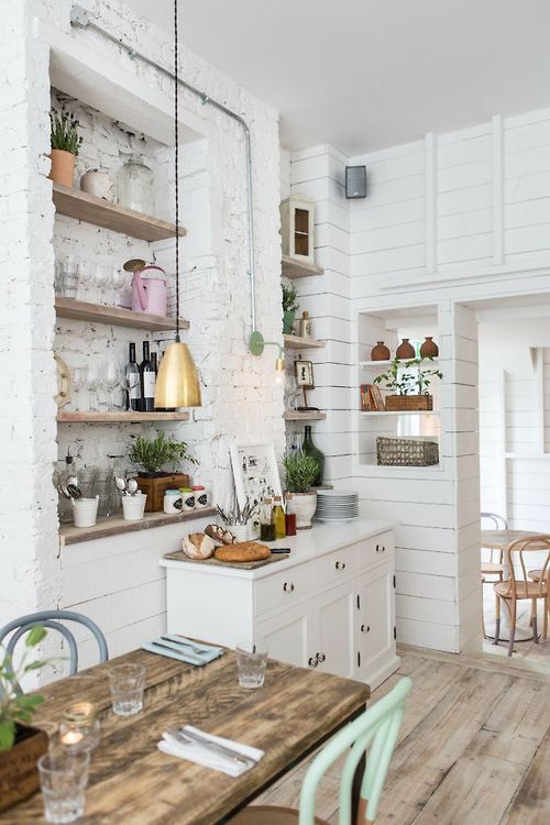 A vintage Scandinavian kitchen with white cabinets, built in shelves, a rough wooden table, pastel chairs and pendant lamps